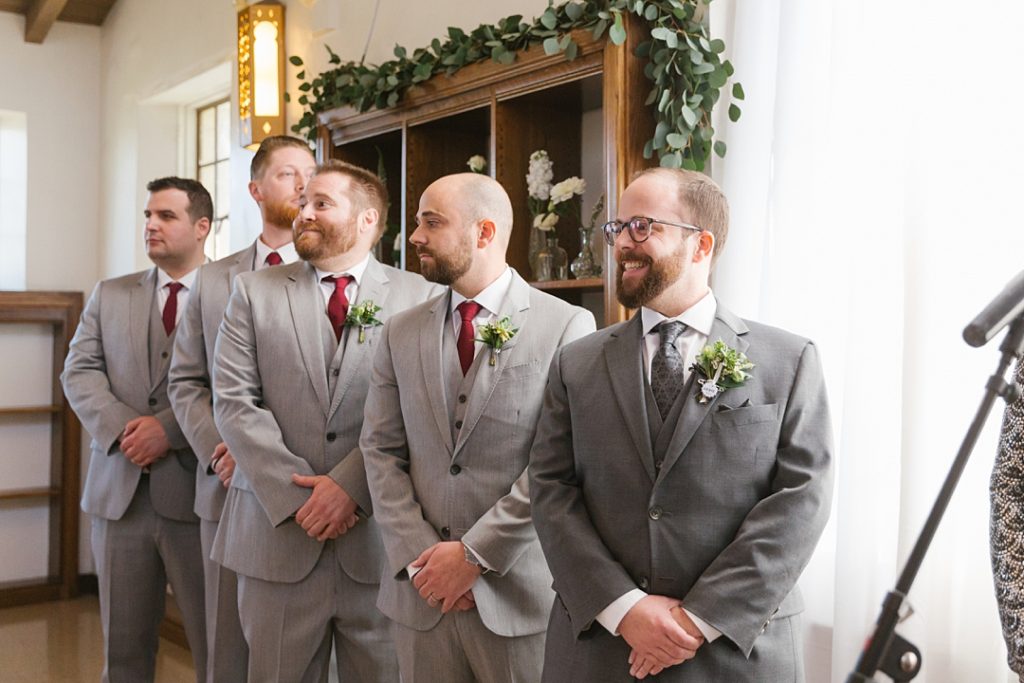 groom in grey suit smiles as bride walks down aisle