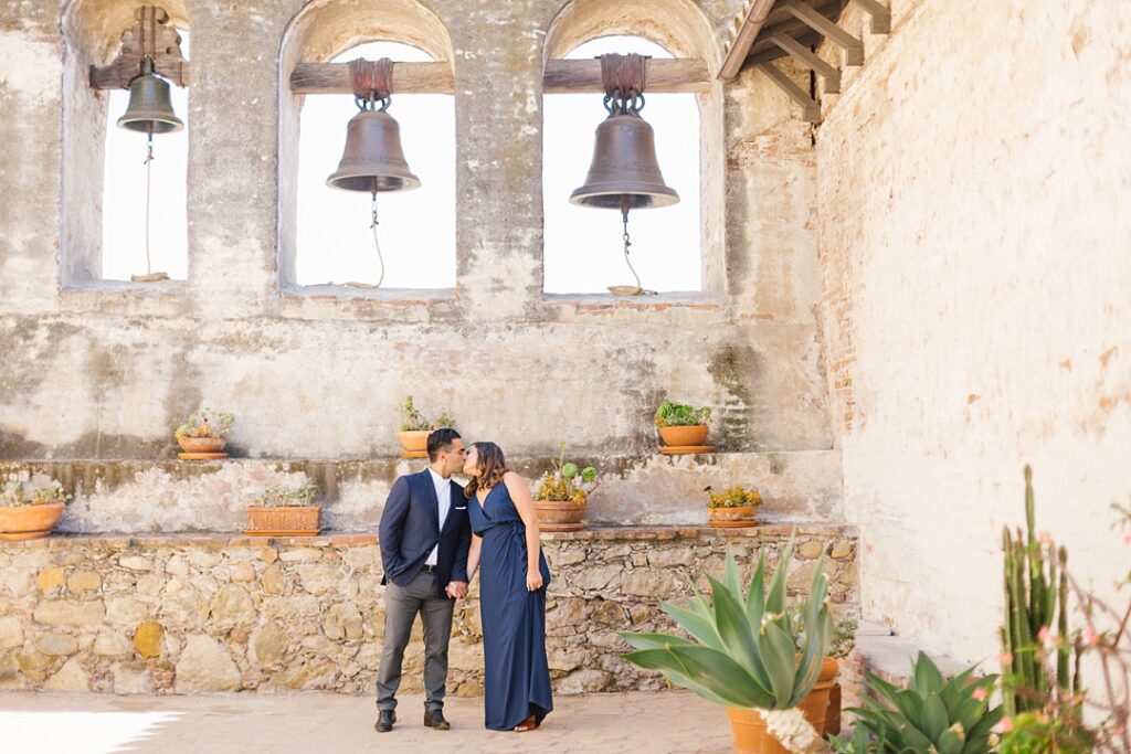 couple is navy blue kisses in front of bells at historic Mission San Juan Capistrano engagement session