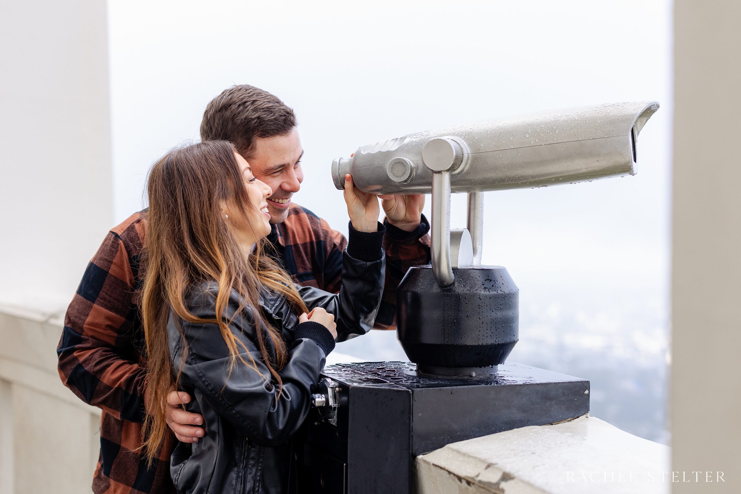 engaged couple looks at views of LA with telescope at Griffith Park